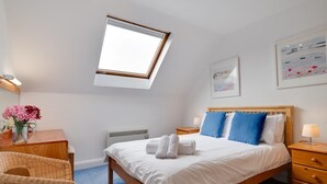 Une chambre avec une lucarne, un lit avec des oreillers bleus et blancs, une table de chevet avec une lampe, une chaise et un tableau encadré au mur.