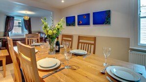A dining area with a wooden table set for four, surrounded by wooden chairs. There is a vase with flowers on the table, and a bottle of wine. The room has a window with curtains, a wall-mounted painting, and a radiator. - 2 Bed in St Ives (oc-pmho08) (Saint Ives)