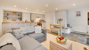 A modern living room with a grey sectional sofa, a dining table with a vase of flowers, and a kitchen in the background. - 1 Bed in St Ives (oc-salt3) (Saint Ives)