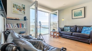 Ein Wohnzimmer mit Ledersofa, Couchtisch aus Holz, Bücherregal, Sofa mit gemusterten Kissen und einem Balkon mit Geländer.