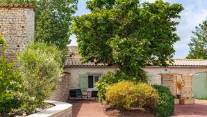 A stone building with a tiled roof, a patio with chairs, and a large tree. - Gîte Le Romarin: Shared Pool, Communal Terrace & Wi-Fi in Courçon (Courçon)