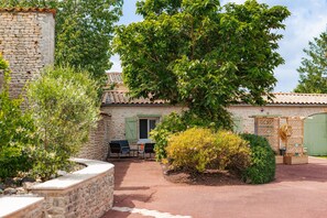 A stone building with a tiled roof, a patio with chairs, and a large tree. - Gîte Le Romarin: Shared Pool, Communal Terrace & Wi-Fi in Courçon (Courçon)