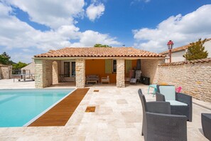Un área junto a la piscina con un patio de piedra, mobiliario exterior y un edificio con techo de tejas.