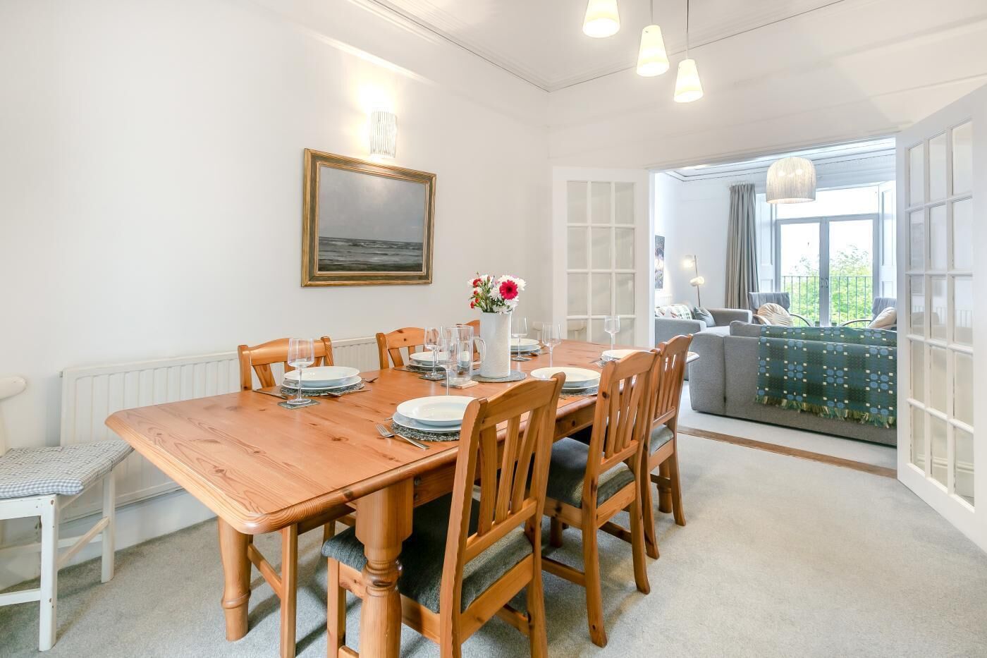 A dining area with a wooden table set for four, a vase with flowers, and a view of the outdoors through a large window.