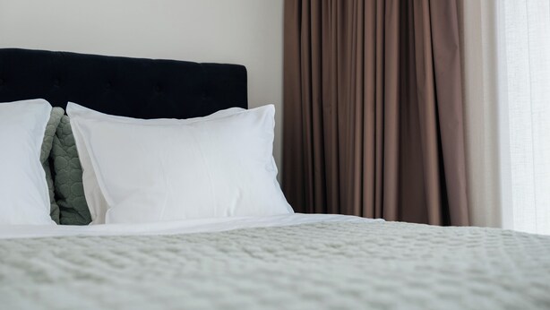 Una cama bien hecha, con almohadas blancas y verdes, cabecera oscura y vistas a cortinas marrones.