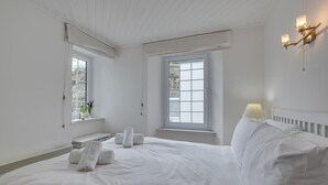 Een slaapkamer in het wit met een groot bed, twee ramen, een nachtkastje en een wandlamp.