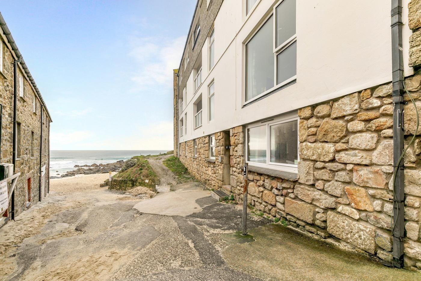Eine schmale Straße mit einem Steinhaus und einem Gebäude mit großen Fenstern. Ein sandiger Pfad führt zum Strand.