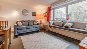 Ein gemütliches Wohnzimmer mit einem grauen Sofa, einem kleinen Tisch, einer Lampe und Blick auf die Stadt durch ein großes Fenster.