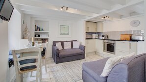 A modern kitchen with white cabinets, a built-in oven, and a microwave. There is a dining area with a white table and chairs, and a grey sofa in the living room. - 2 Bed in St Ives (oc-stg019) (Saint Ives)