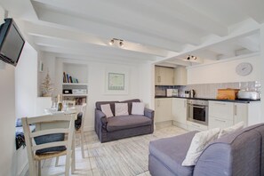 A modern kitchen with white cabinets, a built-in oven, and a microwave. There is a dining area with a white table and chairs, and a grey sofa in the living room. - 2 Bed in St Ives (oc-stg019) (Saint Ives)