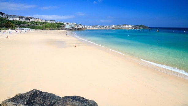 A wide sandy beach with clear blue water, a rocky foreground, and buildings in the background. - 2 Bed in St Ives (oc-stg019) (Saint Ives)