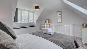A bedroom with a sloped ceiling, a large window with a view of buildings, a bed with a rolled-up towel, and a white dresser. - 2 Bed in St Ives (oc-stg019) (Saint Ives)