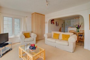 A living room with a TV, two sofas, a coffee table with fruit, and a dining area with a table and chairs. - 1 Bed in St Ives (oc-chyr2) (St Ives)