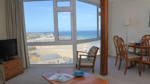 Quarto com uma grande janela com vista para uma praia, uma mesa de madeira com uma tigela e uma televisão sobre um móvel de madeira.