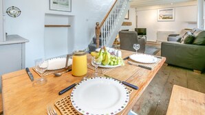 A dining area with a wooden table set for two, featuring a pitcher of orange juice, a plate of fruit, and a glass of white wine. - 2 Bed in St Ives (oc-cotsp) (Saint Ives)