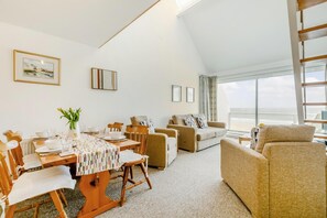 A spacious living area with a dining table set for four, a sofa, and a chair. There are framed pictures on the walls and a view of the sea through large windows. - 1 Bed in St Ives (oc-bpp21) (Saint Ives)
