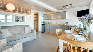 A modern kitchen with stainless steel appliances, a dining area with a wooden table and chairs, and a living room with a sofa and a large window. - 1 Bed in St Ives (oc-crab01) (St Ives)