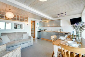 A modern kitchen with stainless steel appliances, a dining area with a wooden table and chairs, and a living room with a sofa and a large window. - 1 Bed in St Ives (oc-crab01) (St Ives)