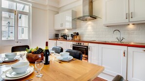 A modern kitchen with white cabinetry, a wooden dining table set for four, and a large window providing natural light. - 2 Bed in St Ives (oc-clv2a) (Saint Ives)