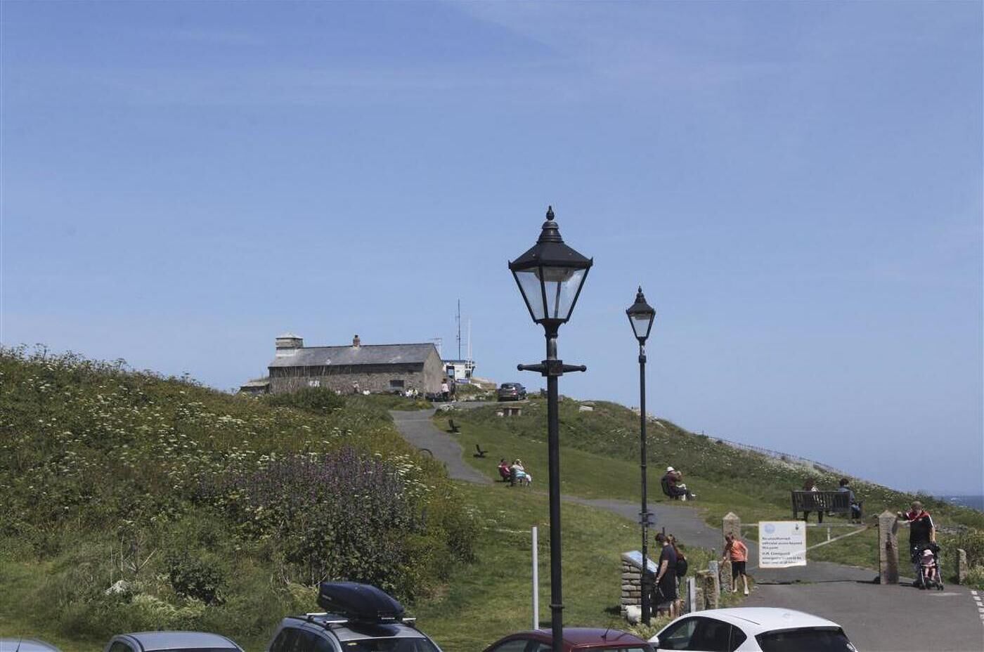 A coastal path with a stone building in the background, a paved walkway, and a few people walking.