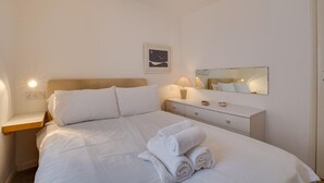 Ein ordentlich bezogenes Bett mit weißen Leinentüchern und aufgerollten Handtüchern, ein Nachttisch mit einer Lampe, ein kleines gerahmtes Bild an der Wand und ein Spiegel, der das Bett reflektiert.