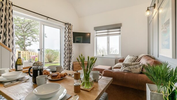 A cozy living room with a wooden dining table set for breakfast, a brown leather sofa, and a large window with curtains. - 2 Bed in St Ives (oc-godct7) (Carbis Bay)
