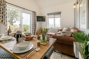 Un salon chaleureux avec une table à manger en bois, préparée pour le petit-déjeuner, un canapé en cuir marron et une grande fenêtre avec des rideaux.
