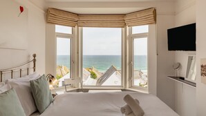 Un dormitorio con cama, mesita de noche, lámpara, ventana con vistas al mar y televisor de pared.