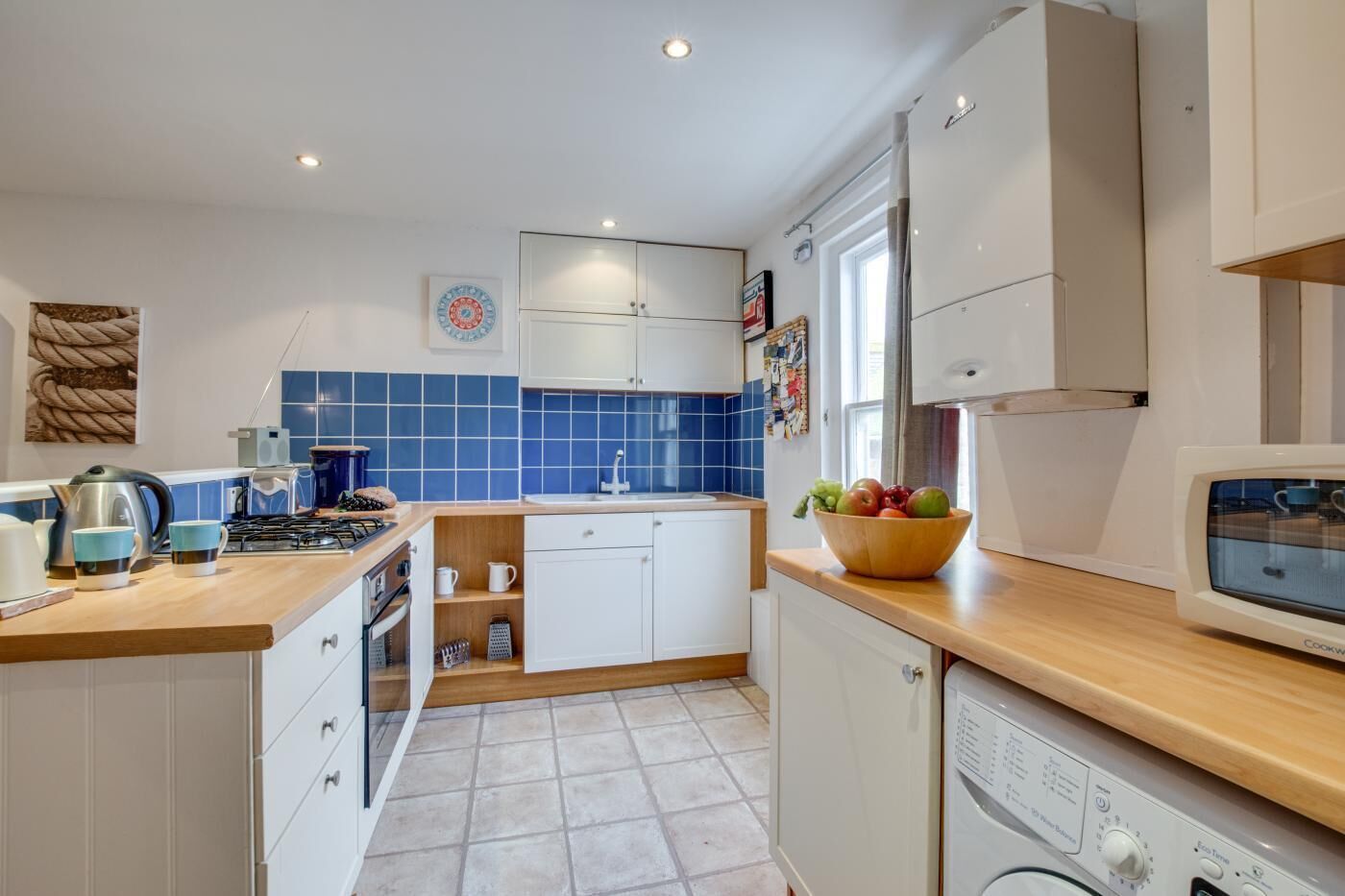 Una cocina moderna con armarios blancos, un respuesto de azulejos azules, una encimera de madera, una estufa a gas, un microondas y un tazón de frutas.