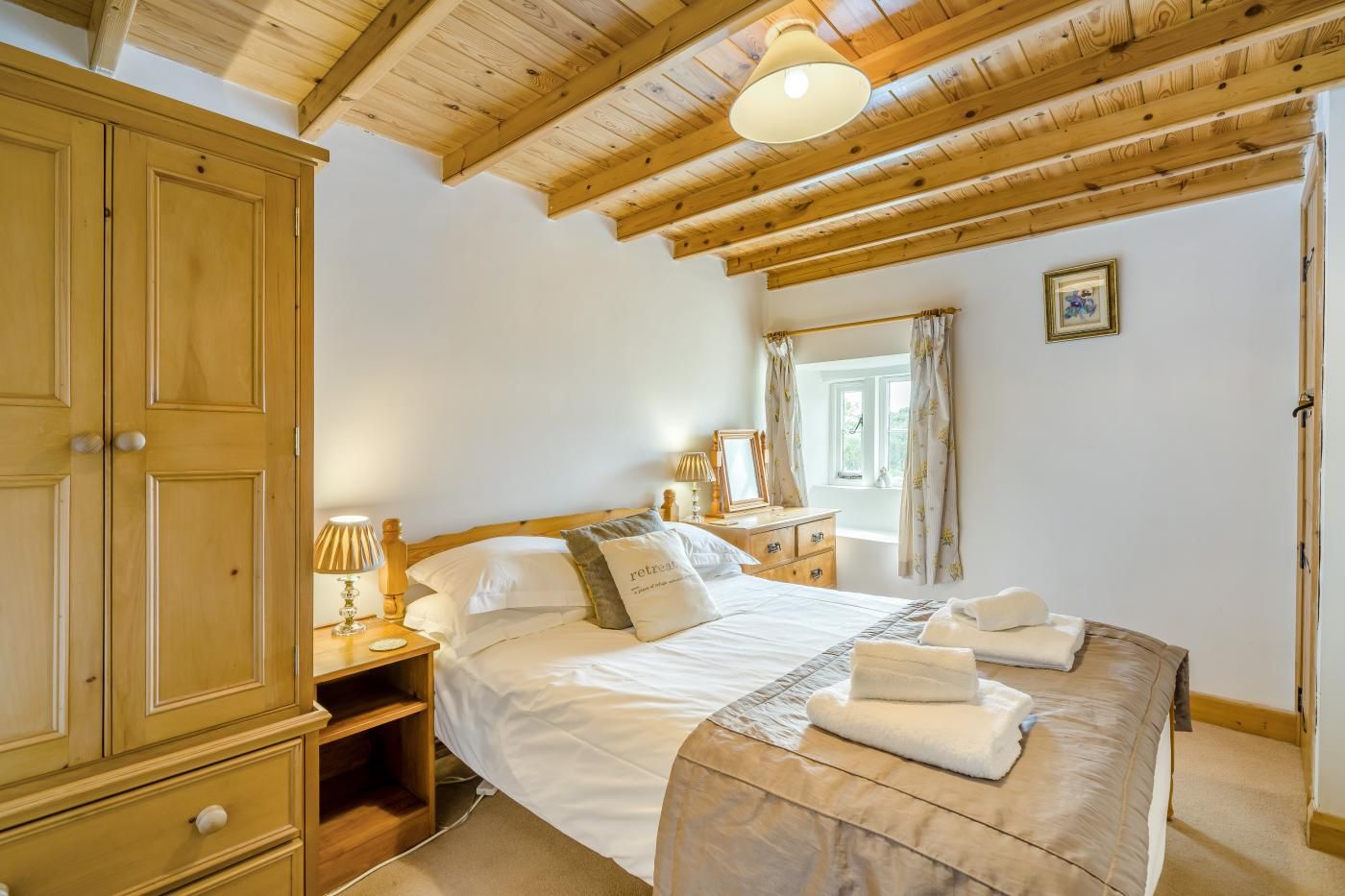 Un dormitorio con un armario de madera, una cama con ropa de cama blanca, una mesita de noche y una lámpara en la mesita auxiliar, además de una ventana con cortinas.