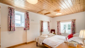 Ein Schlafzimmer mit Holzboden, zwei Fenstern mit blumigen Vorhängen, einem Bett mit weißen Leinen, einem Nachttisch mit Lampe und einem roten Kissen.