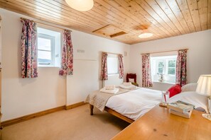 Ein Schlafzimmer mit Holzboden, zwei Fenstern mit blumigen Vorhängen, einem Bett mit weißen Leinen, einem Nachttisch mit Lampe und einem roten Kissen.