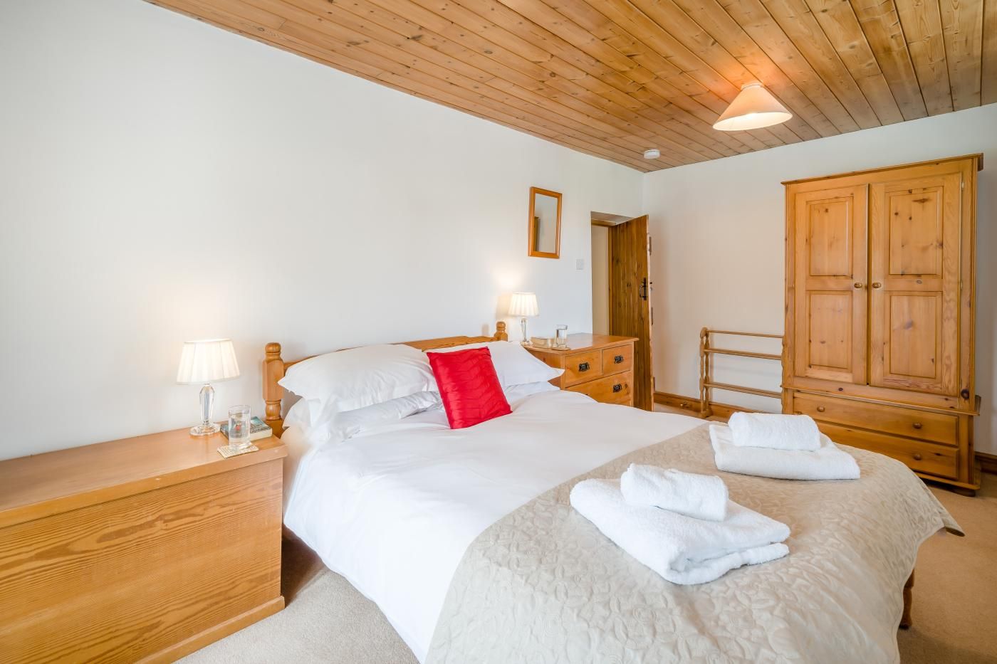 A hotel room with a wooden ceiling, a bed with white bedding and a red pillow, a wooden wardrobe, and a nightstand with a lamp.