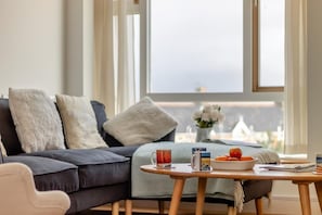 A modern living room with a dark sofa, a round wooden table with a bowl of fruit, a mug, and a book. - 2 Bed in Southwold (oc-tibw) (Southwold)