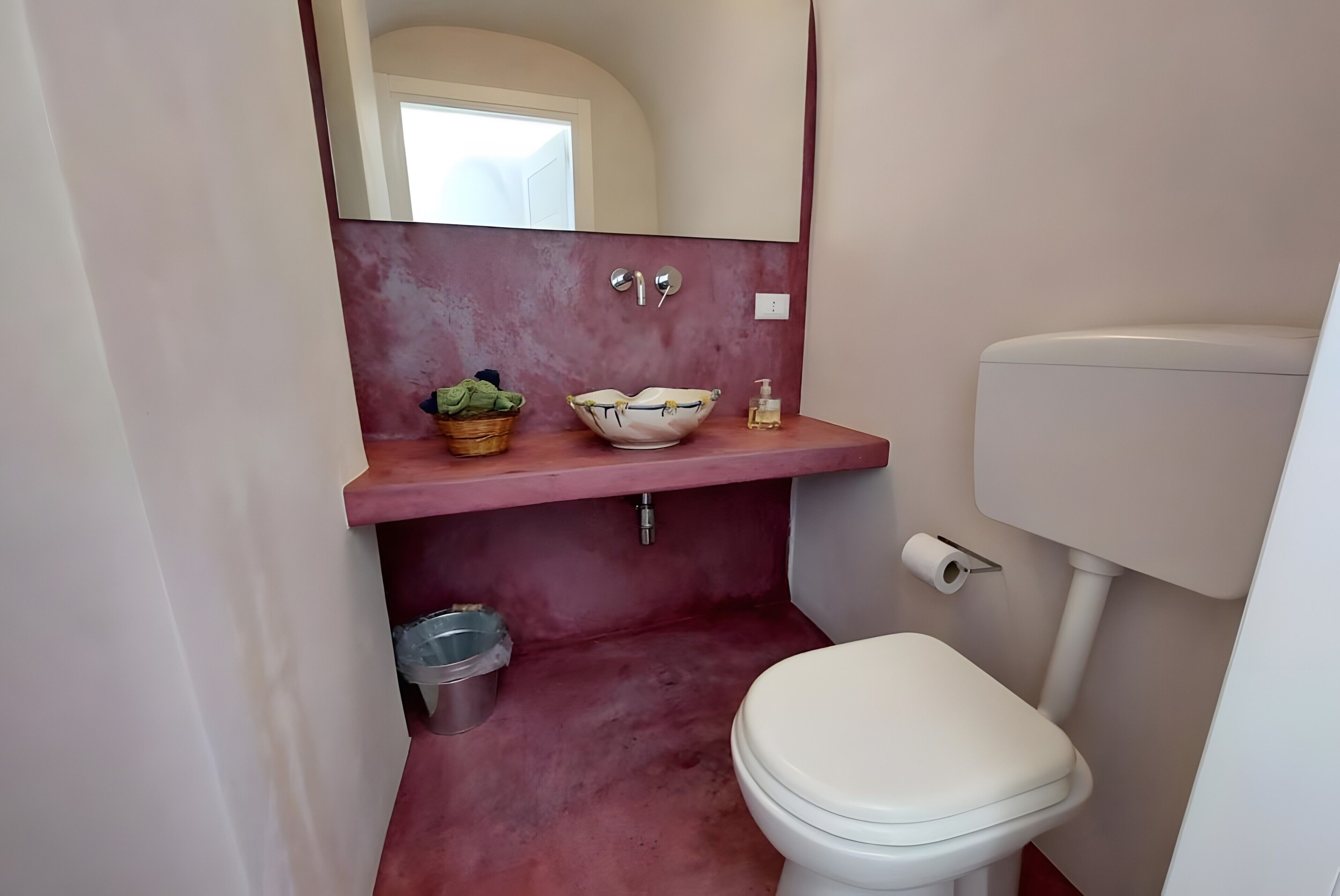 Une salle de bain avec des toilettes blanches, un lavabo rose avec un receveur, un miroir et une petite poubelle.