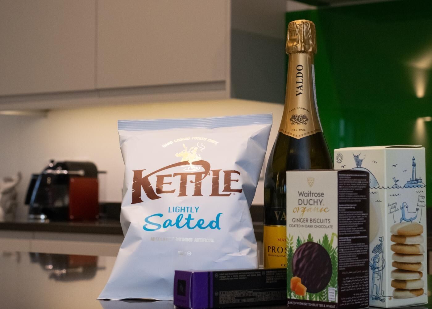 Una busta di chips Kettle leggermente salate, una bottiglia di champagne Valdo, biscotti allo zenzero biologici Waitrose Duchy e una scatola di biscotti.