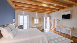 Een slaapkamer met een groot bed, houten balken in het plafond, een bureau en een aan de muur bevestigde televisie.