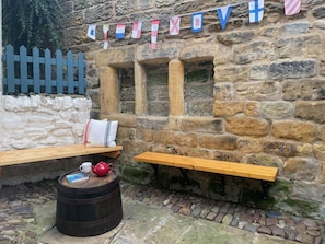 A stone wall with wooden benches and a barrel, a flag with flags, and a white picket fence.