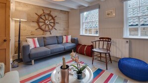 A cozy room with a grey sofa, a wooden chair, a ship's wheel wall decoration, a large window with patterned glass, and a colorful striped rug. - 3 Bed in Robin Hoods Bay (oc-r041) (Robin Hood's Bay)