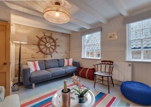 Ein gemütlicher Raum mit einem grauen Sofa, einem Holzstuhl, einer Wanddekoration mit Schiffsschwengel, einem großen Fenster mit Buntglas und einem farbenfrohen Streifen Teppich.