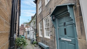 A narrow street with stone buildings, a green door labeled 'FENAY COTTAGE,' and a small garden area with a bench and potted plants. - 3 Bed in Robin Hoods Bay (oc-r041) (Robin Hood's Bay)