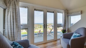 Ein Zimmer mit einer großen Schiebeglastür, die auf einen Balkon mit Meerblick führt. Es gibt zwei graue Sessel mit blauen Kissen mit Muster.