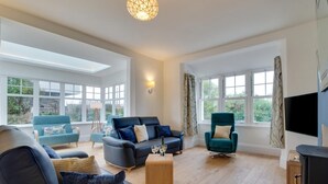 Ein geräumiges Wohnzimmer mit einer großen Couch, einem kleineren Sessel und einem türkisblauen Sessel. Ein Holztisch steht davor, mit einer Flasche und einem Glas darauf. Das Zimmer verfügt über große Fenster mit Vorhängen, einen Flachbildfernseher und einen Kamin.