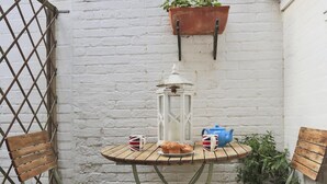 Een klein terras met een ronde tafel, twee houten stoelen, een hangende bloemenplanter, een lantaarn en een theepot.
