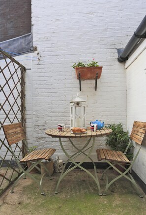 A small outdoor seating area with a round table, two wooden chairs, a hanging planter with flowers, a lantern, and a teapot. - 2 Bed in Southwold (oc-aatrin) (Southwold)