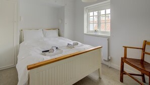 Ein helles Schlafzimmer mit einem hölzernen Bettgestell, einem weißen Heizkörper, einem Fenster mit Blick auf Gebäude und einem Holzstuhl.