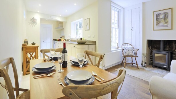 Un comedor con mesa de madera dispuesta para cuatro, una chimenea y una cocina al fondo.