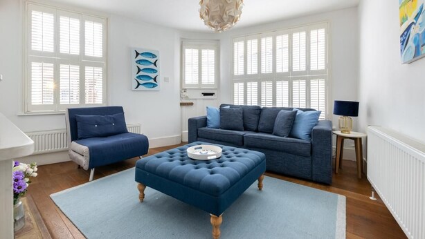Ein modernes Wohnzimmer mit einem blauen Sofa, einem blauen Sessel und einem gepolsterten Hocker. Große Fenster mit Fensterläden, ein weißer Heizkörper und eine dekorative Deckenleuchte sind ebenfalls zu sehen.