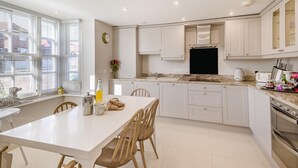 A modern kitchen with white cabinets, a dining table, and chairs. There is a window with blinds, a clock, and a vase with flowers.