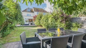 A patio with wicker furniture and a glass table, surrounded by greenery and a house in the background.